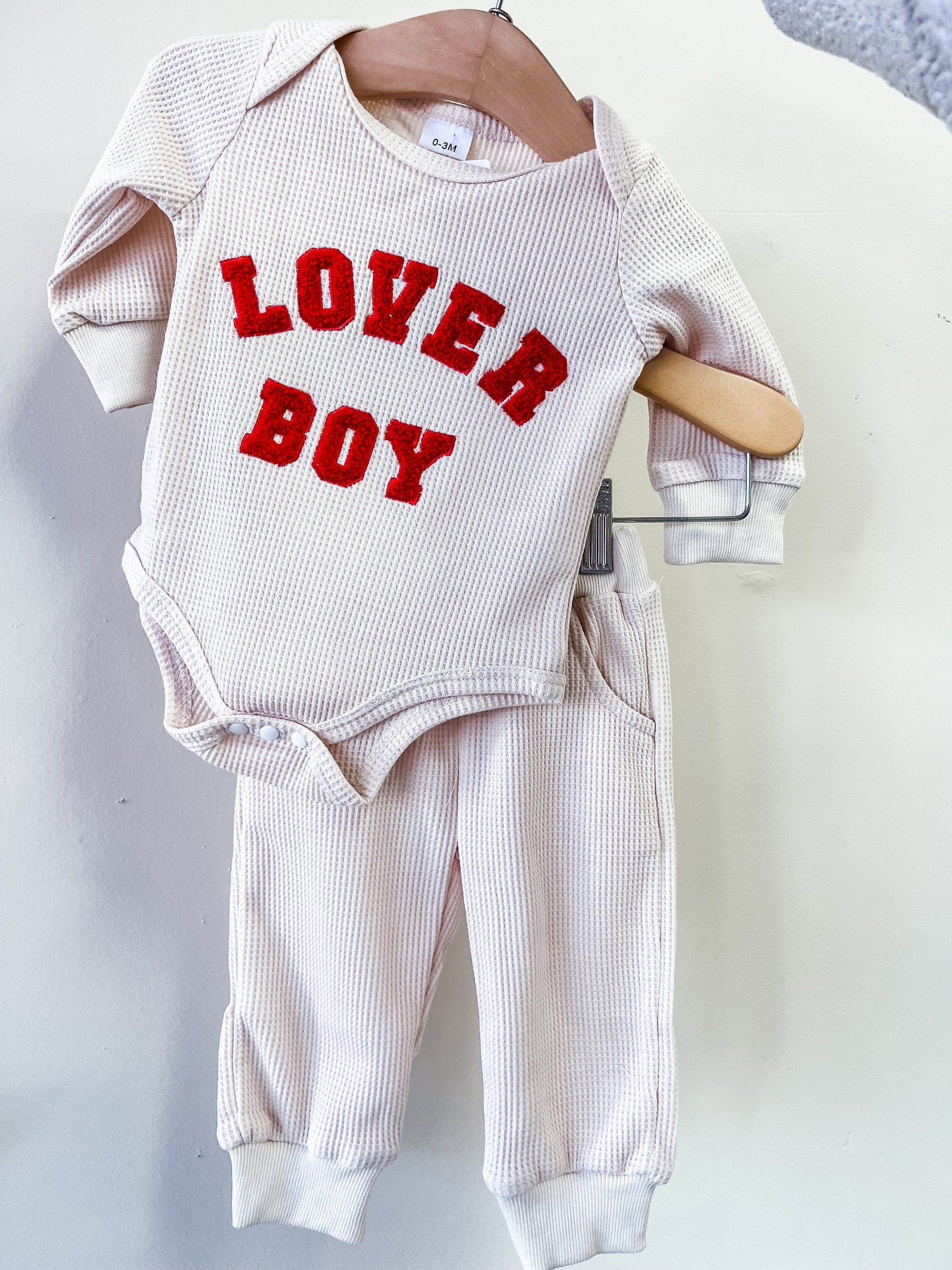 The Loved Boy Waffle Set by Tiny Tot Bundles features a cream long-sleeve onesie with red "LOVED BOY" lettering and matching pants, both displayed on a wooden hanger against a white background.