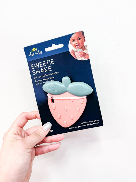 A hand holds up an itzy ritzy Strawberry Sweetie Teether w/ Rattle in packaging featuring a baby and noting it soothes sore gums.