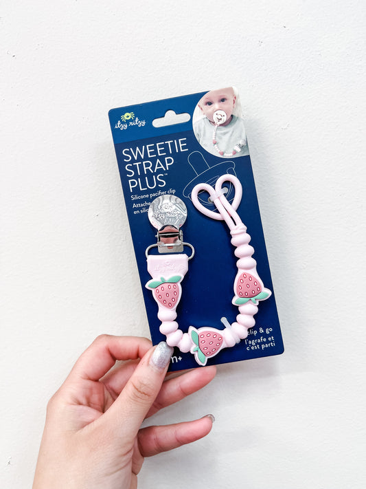 A hand holds an itzy ritzy Strawberry Sweetie Pacifier Clip featuring pink beads and strawberry designs, packaged and displayed against a white background.