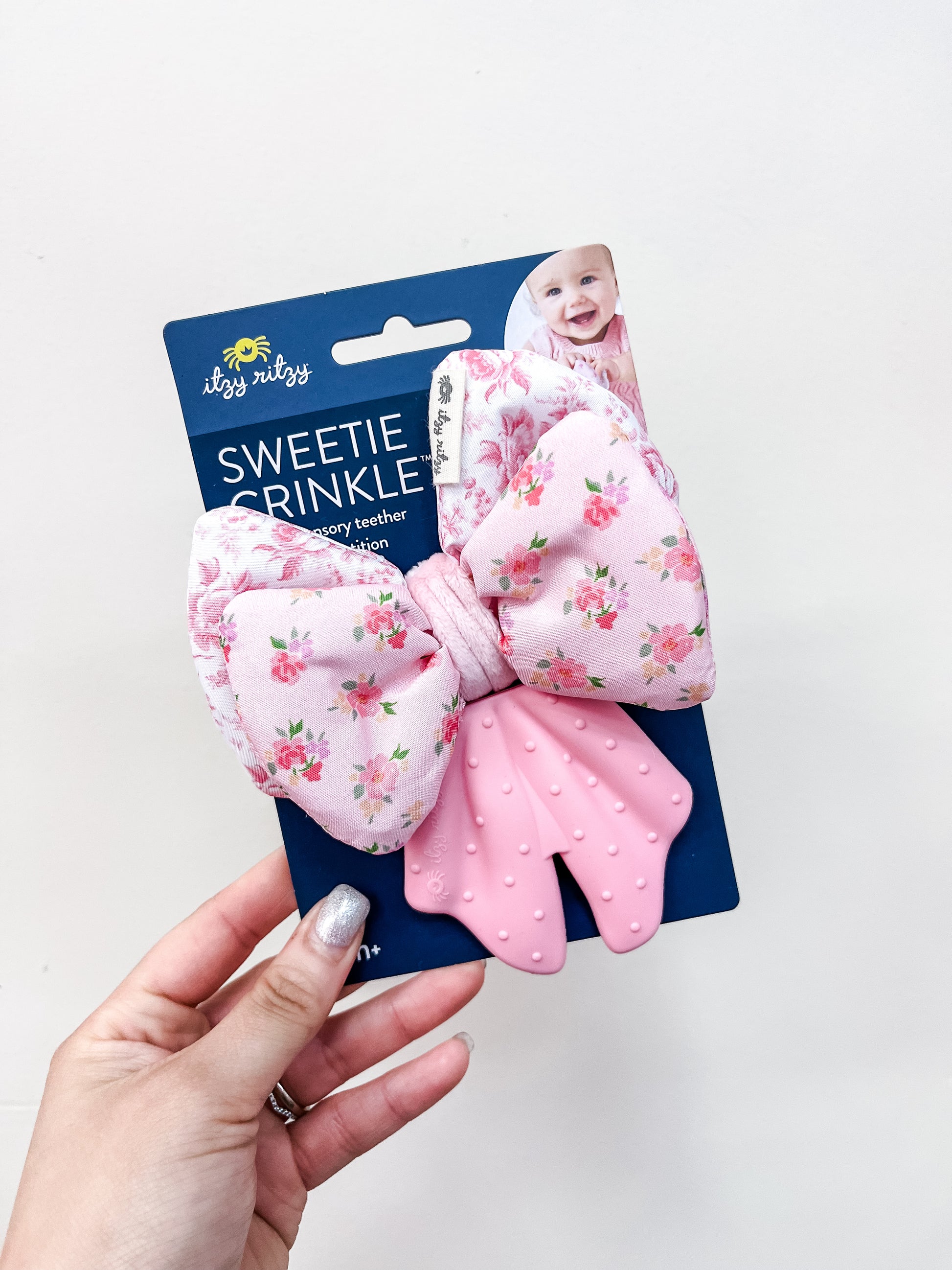 A hand holds an itzy ritzy Sweetie Crinkle Bow Teether package, featuring a pink floral bow and mitten-shaped textured pink teether. The packaging displays a smiling baby in the corner.