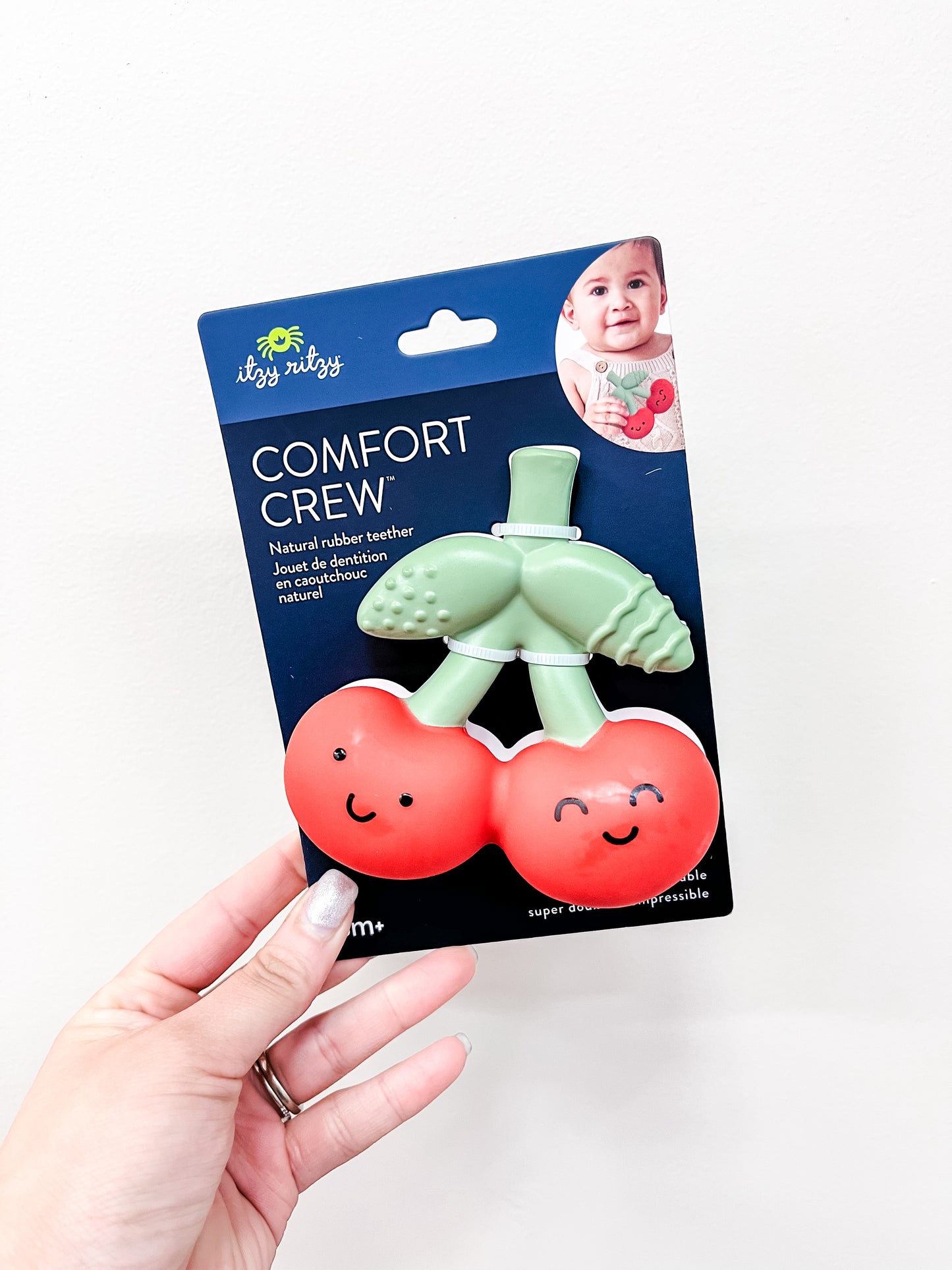 A hand holds an itzy ritzy Happy Cherries Teething Toy made of natural rubber, shaped like smiling cherries with green leaves. The blue package displays a baby’s photo in the top right corner.