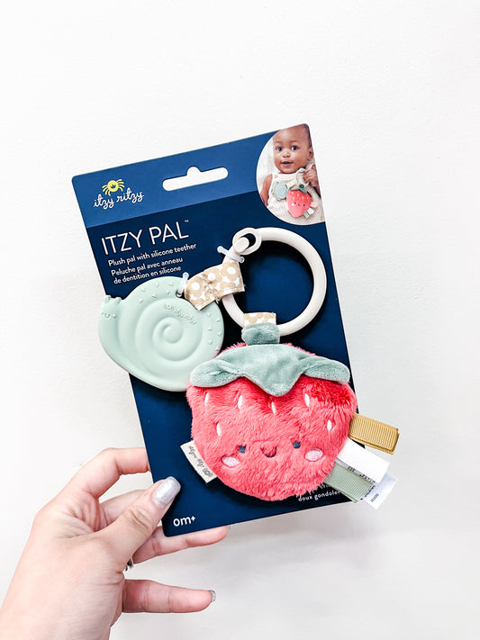 A hand holds the itzy ritzy Berry Friends Sensory Teether, a baby toy for ages 0+ featuring a plush strawberry character, mint green silicone teether, and fabric loop.