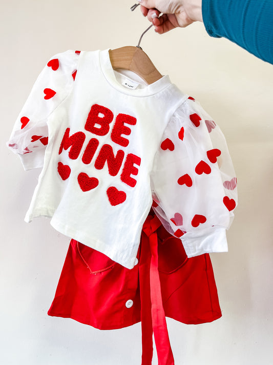 A hand holds up the Tiny Tot Bundles Sweetheart Puff Skirt Set, featuring a white top with red "BE MINE" and heart puffy sleeves, paired with a red skirt with bow and pockets.