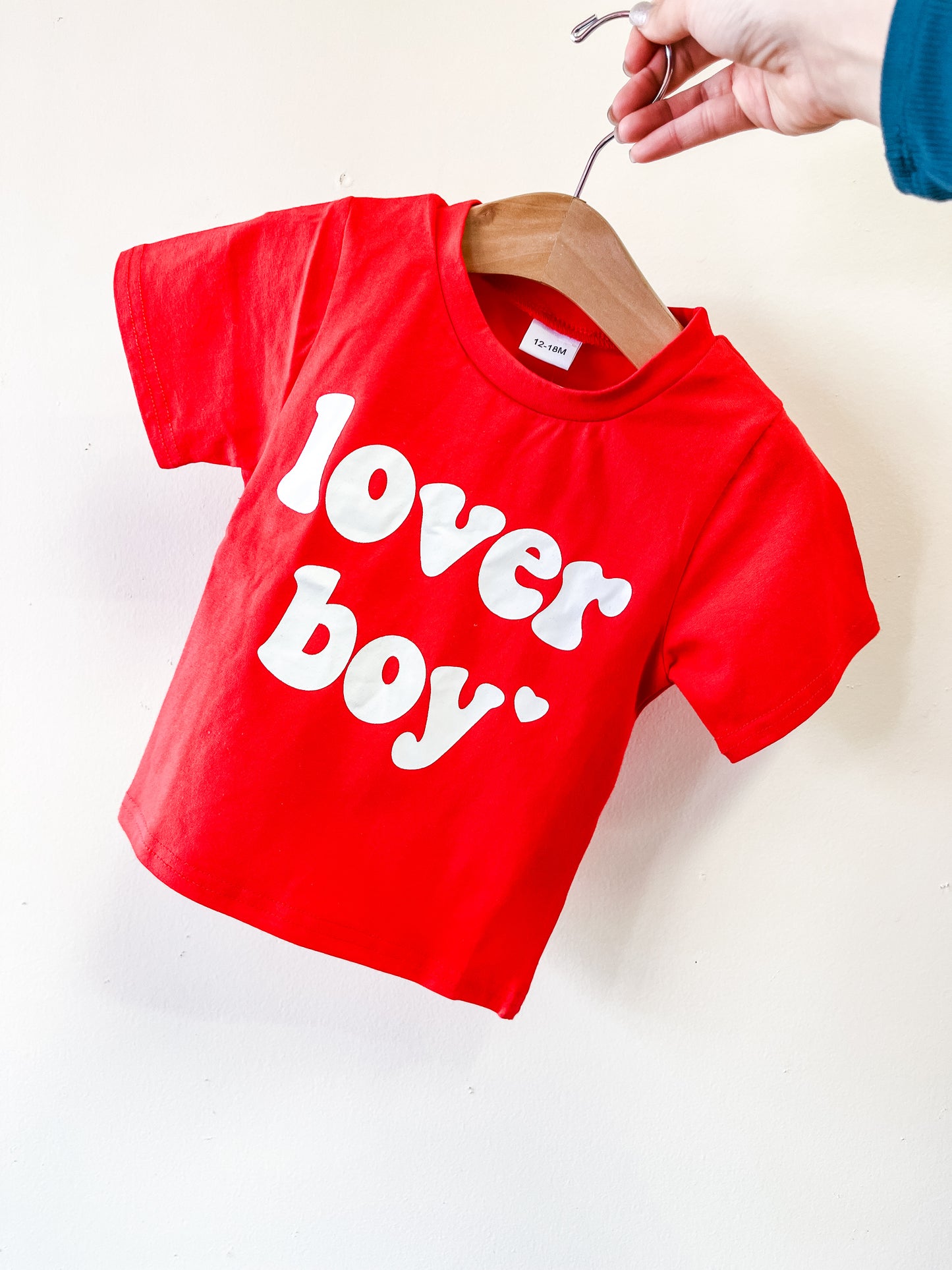 A hand displays the red Lover Boy Tee by Tiny Tot Bundles, toddler-sized and featuring bold white "lover boy" lettering on the front, hanging on a wooden hanger against an off-white wall.