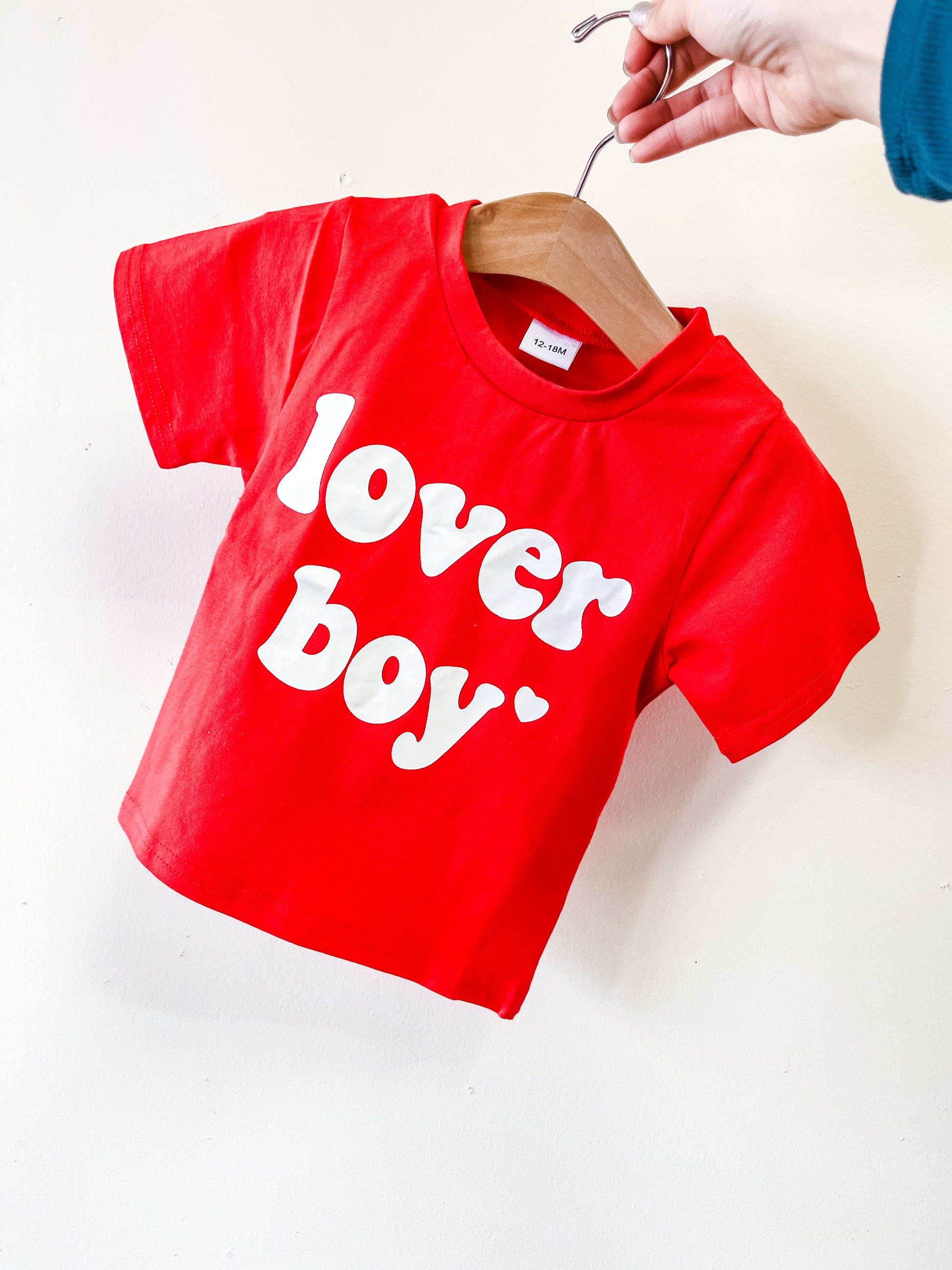 A hand displays the red Lover Boy Tee by Tiny Tot Bundles, toddler-sized and featuring bold white "lover boy" lettering on the front, hanging on a wooden hanger against an off-white wall.