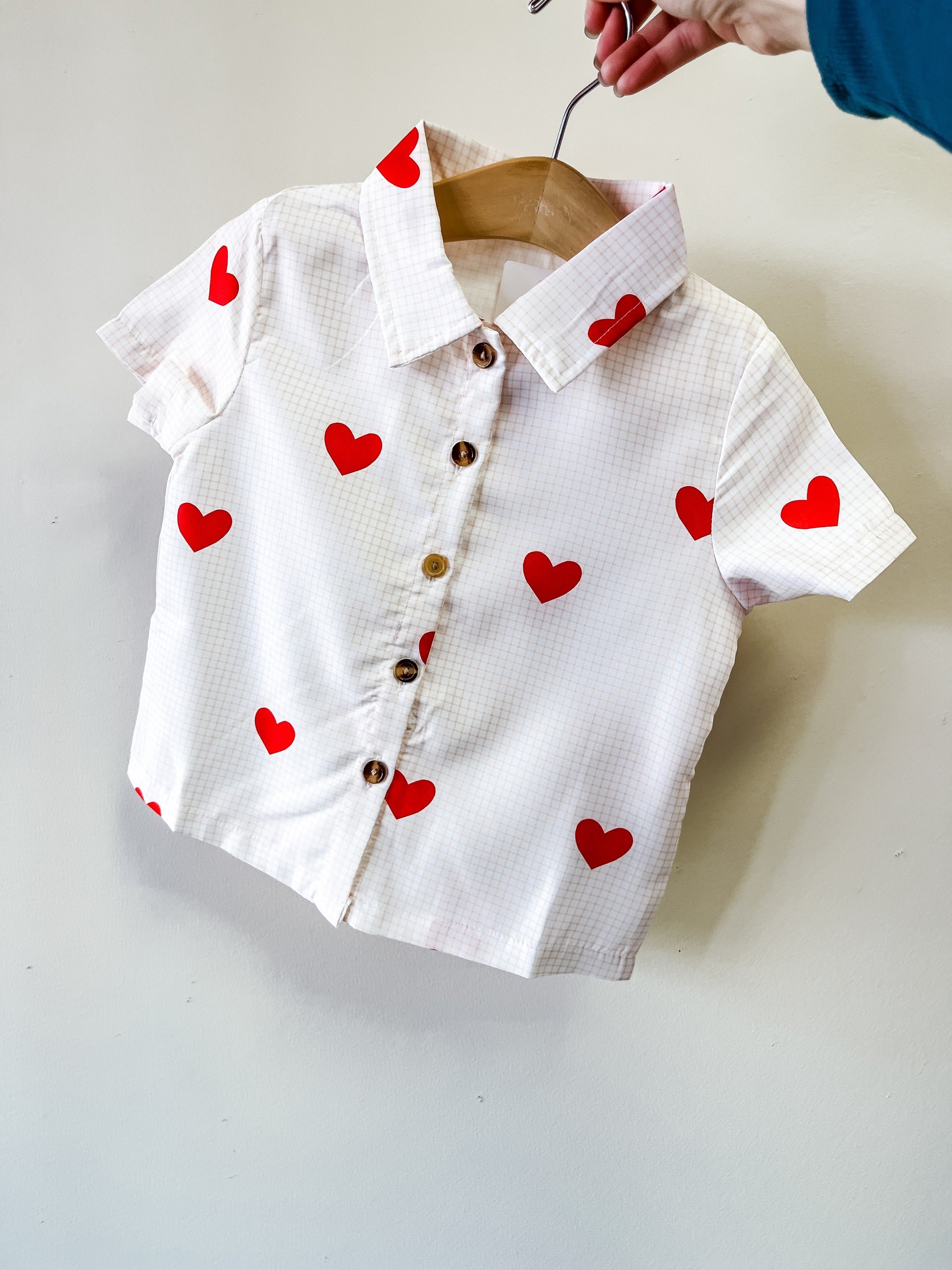 A hand holds the Sweetheart Button-Up Shirt by Tiny Tot Bundles—a white, short-sleeve children's shirt with a subtle checkered pattern and red heart prints, displayed on a hanger.