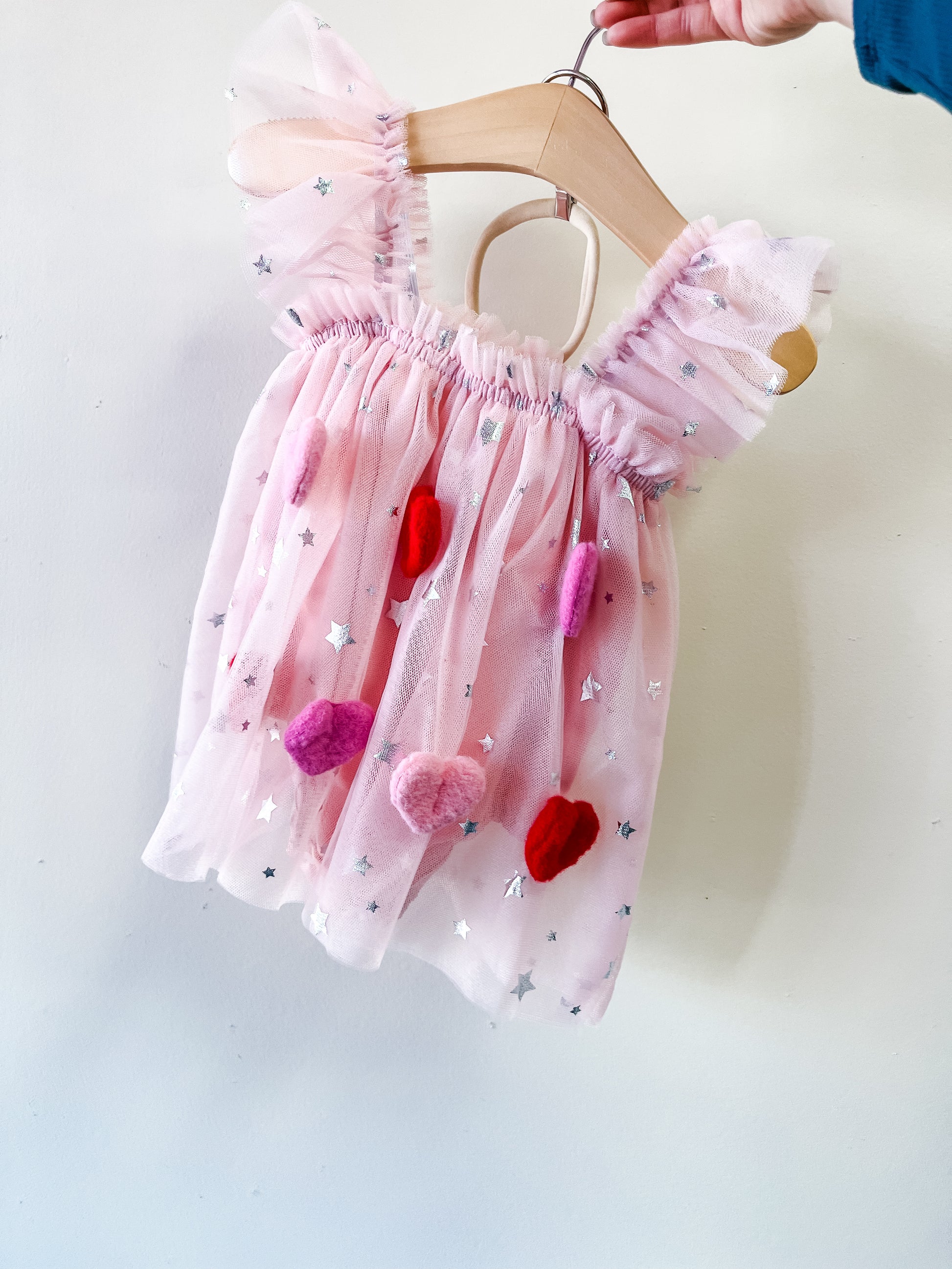 The Starry Sweetheart Tulle Dress by Tiny Tot Bundles is a pink toddler dress with silver star accents and colorful pom-poms, shown on a wooden hanger against a plain light background.