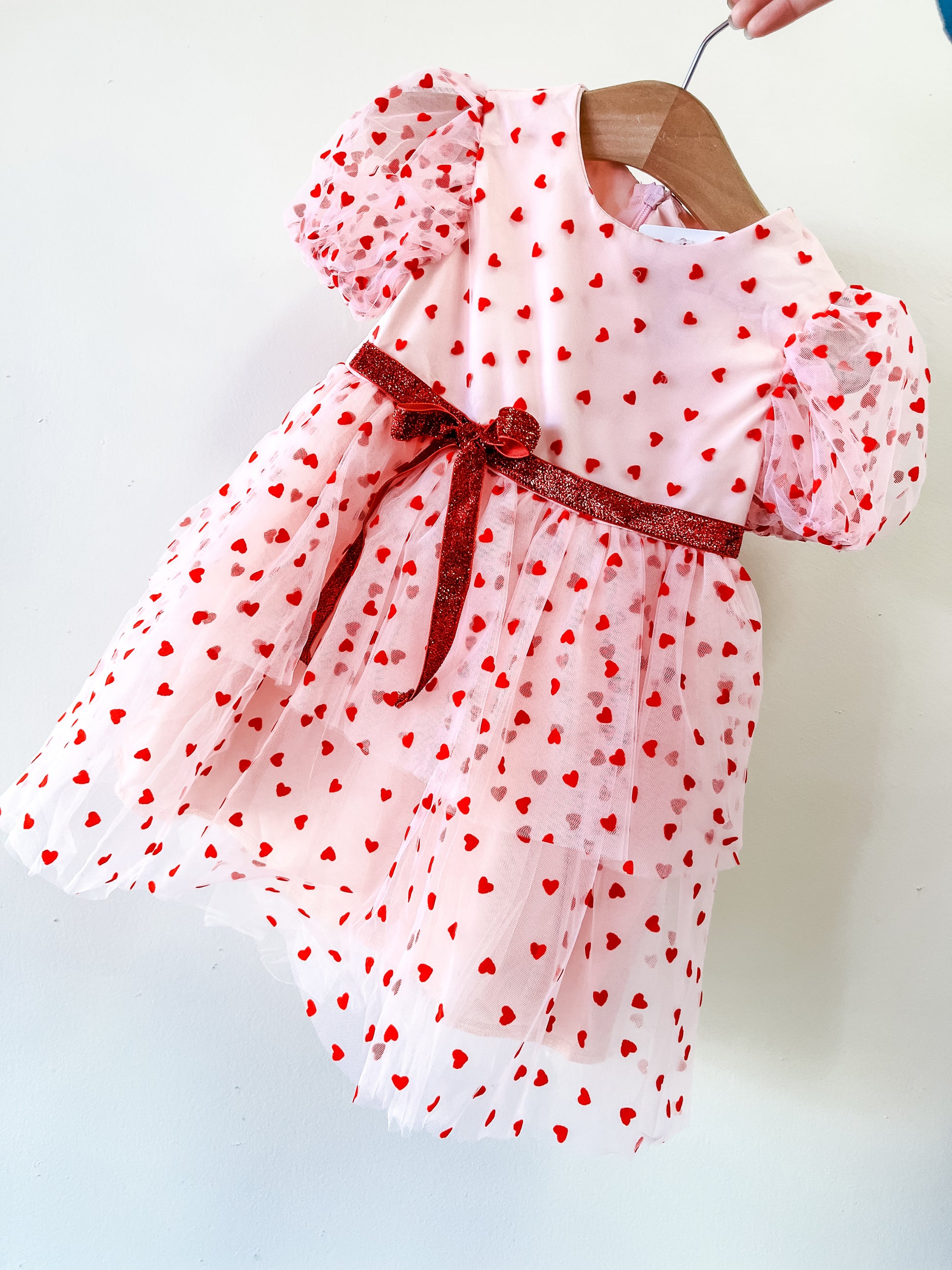 The Valentine's Tulle Dress by Tiny Tot Bundles is a pink toddler dress with red heart patterns, short puffy sleeves, and a red glittery waist ribbon, shown on a wooden hanger against a white background.