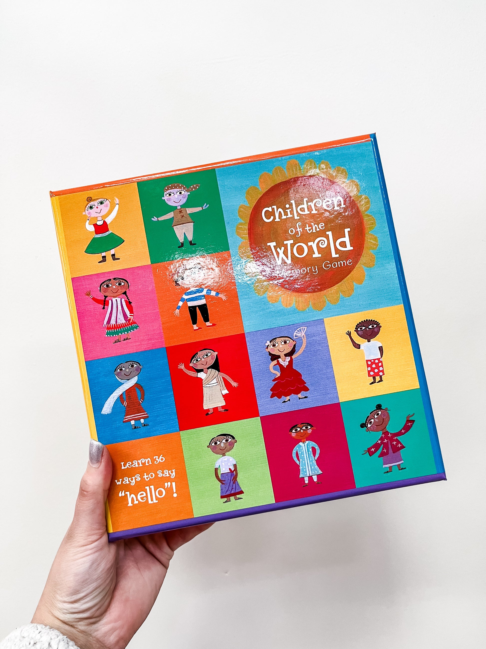 A hand holds Barefoot Books’ Children of the World Memory Game, showing illustrated children in traditional dress from many cultures on a grid. Bottom text says, “Learn 36 ways to say hello!”.
