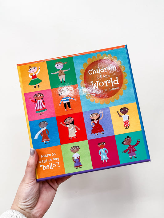 A hand holds Barefoot Books’ Children of the World Memory Game, showing illustrated children in traditional dress from many cultures on a grid. Bottom text says, “Learn 36 ways to say hello!”.