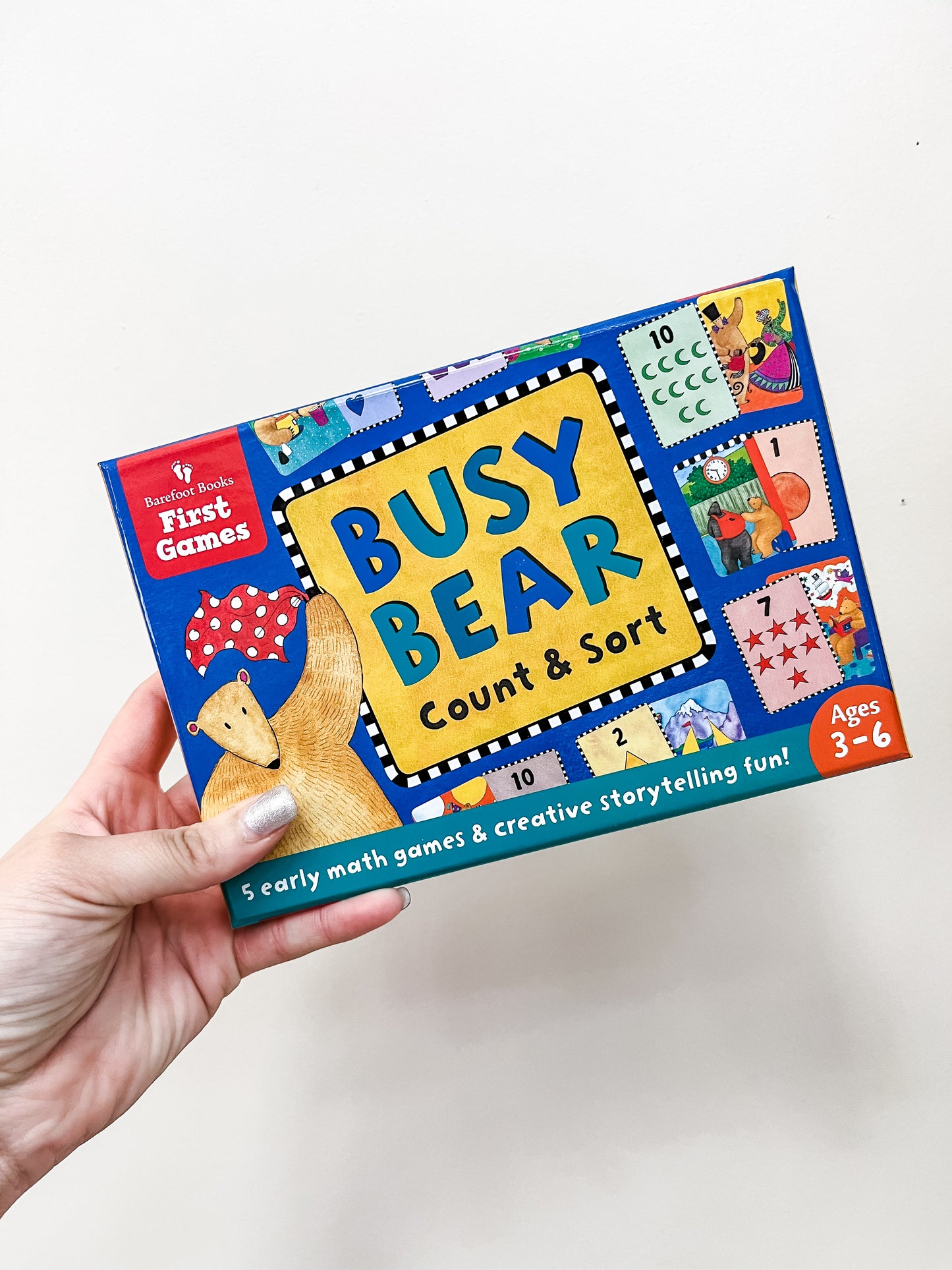 A hand holds a box of Barefoot Books' Busy Bear Count and Sort Game, a blue box with colorful numbers, bear illustrations, and the tagline: 5 early math games & creative storytelling fun! For ages 3–6.