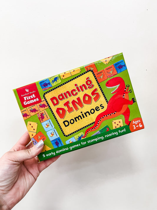 A hand holds a vibrant Dancing Dinos Dominoes Game box by Barefoot Books, showing a red dino and domino illustrations. Designed for ages 3-6, it includes 5 early domino games for children.