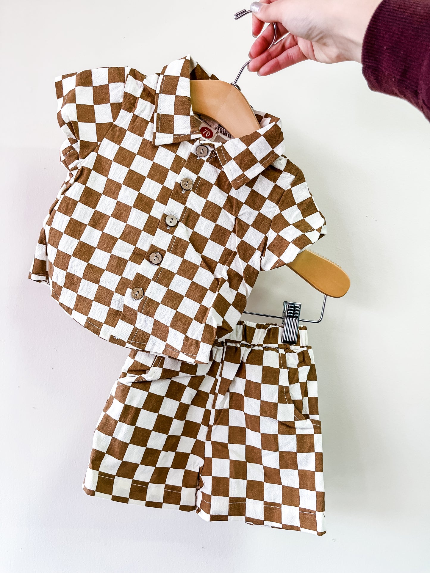 A hand holds the Tiny Tot Bundles Mocha Check Mate Set, a matching short-sleeve button-up shirt and shorts with a brown and white checkerboard pattern, displayed on a hanger against a plain white background.