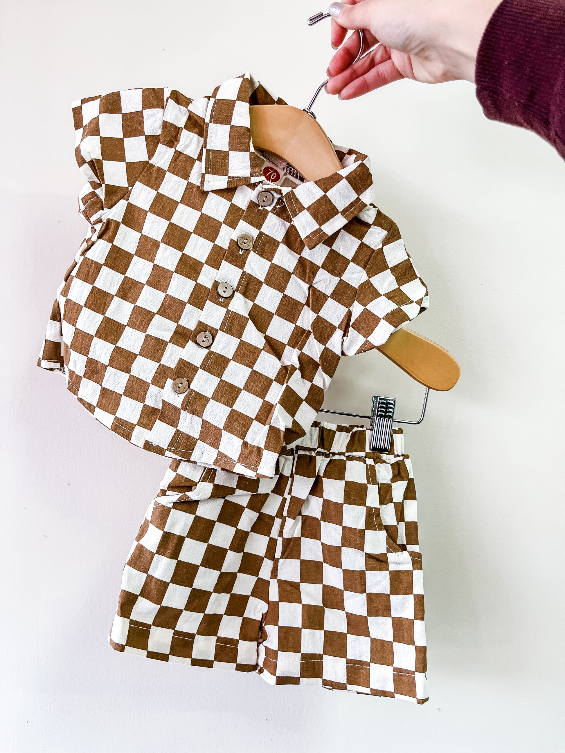 A hand holds the Tiny Tot Bundles Mocha Check Mate Set, a matching short-sleeve button-up shirt and shorts with a brown and white checkerboard pattern, displayed on a hanger against a plain white background.