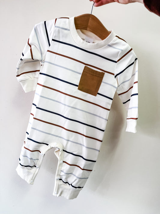 A hand displays Tiny Tot Bundles’ Neutral Stripe Pocket Romper, a white long-sleeve baby outfit with horizontal navy, brown, and orange stripes and a brown square chest pocket.