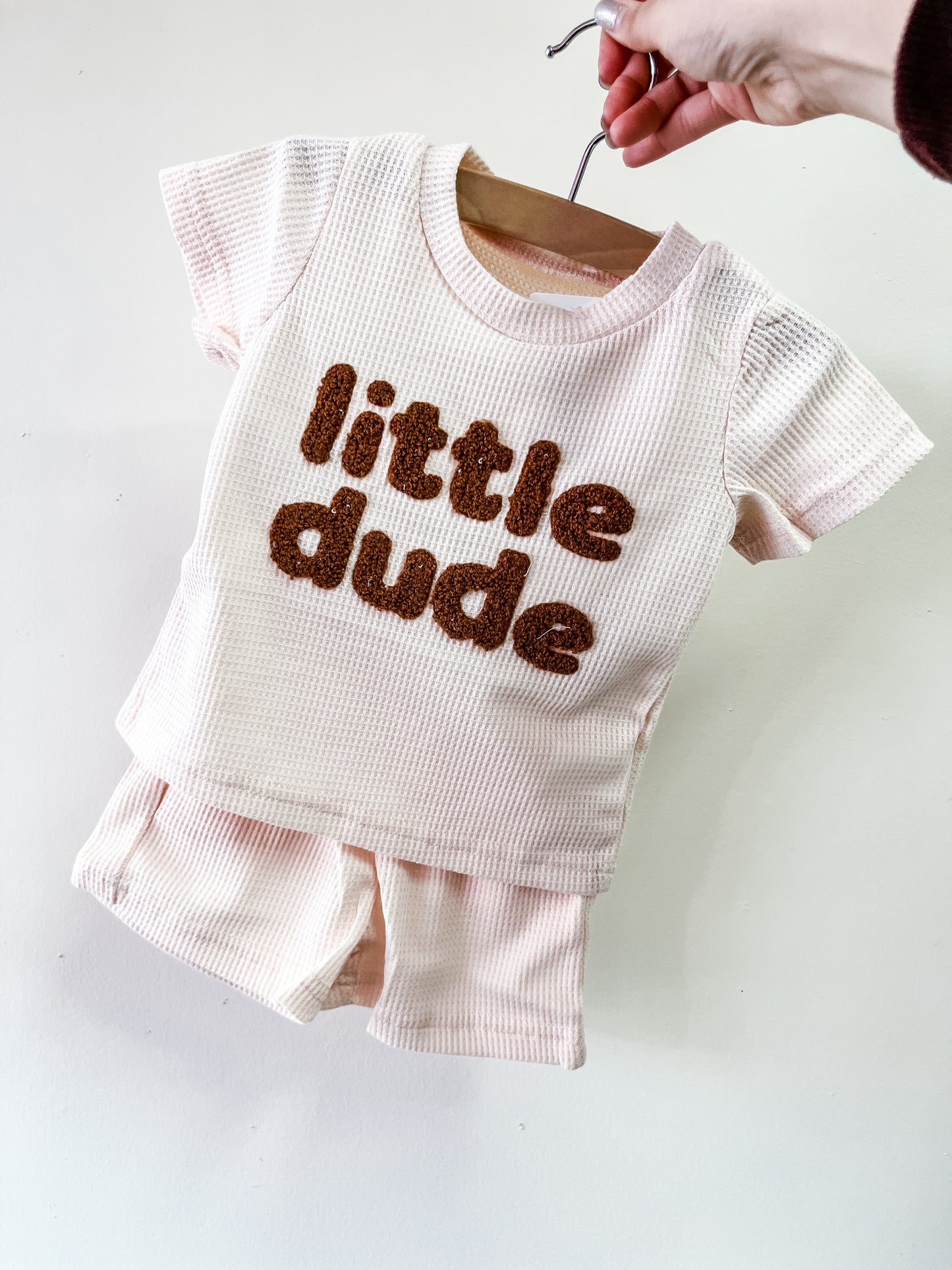 A hand holds a hanger with Tiny Tot Bundles' Little Dude Waffle Set, featuring a light pink, short-sleeved textured shirt with "little dude" in bold brown fuzzy letters and matching shorts underneath.