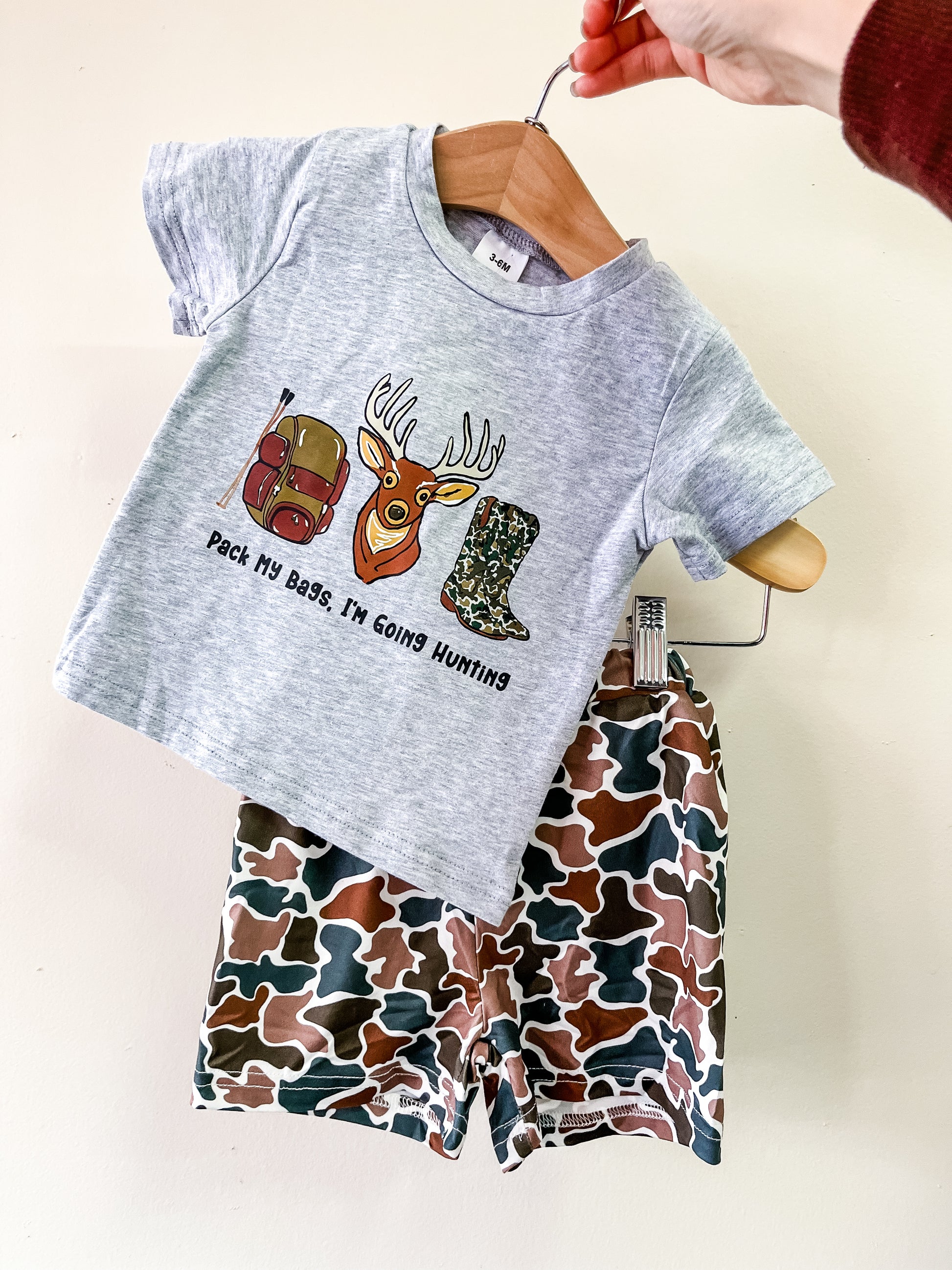 A hand displays the Tiny Tot Bundles Pack My Bags Hunting Set, featuring a gray t-shirt with a backpack, deer head, boot, and “Pack My Bags I’m Going Hunting” text, paired with brown, green, and tan camo shorts.