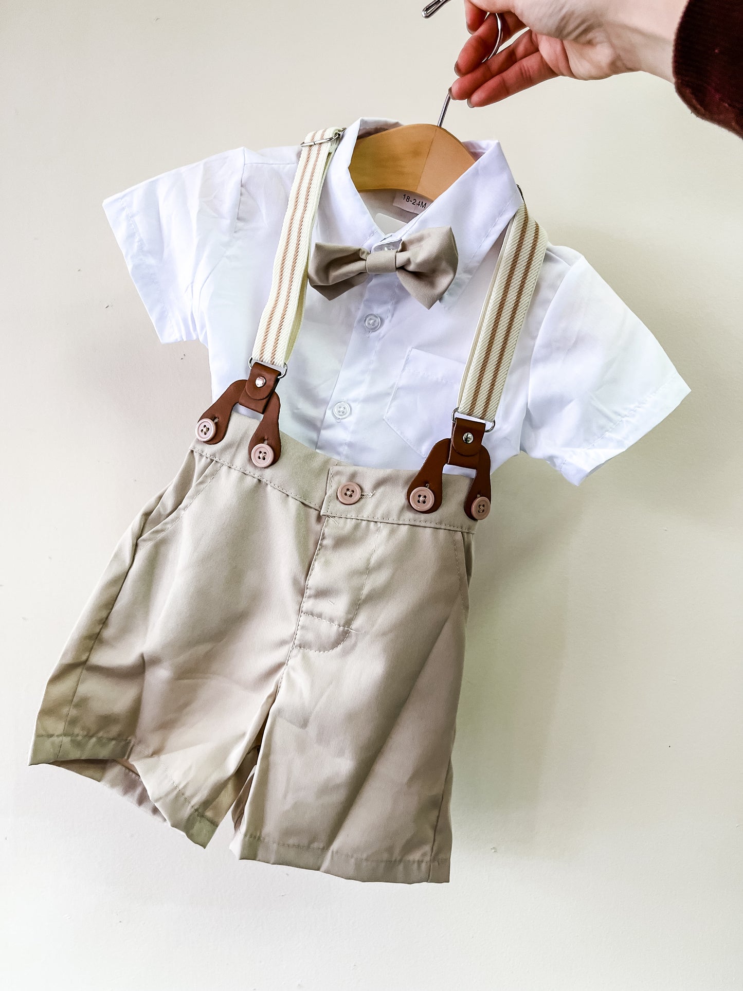 A hand holds the Classic Bowtie Suspender Set by Tiny Tot Bundles: a child's white short-sleeve shirt, beige bow tie, beige shorts, and matching suspenders on a hanger against a light background.