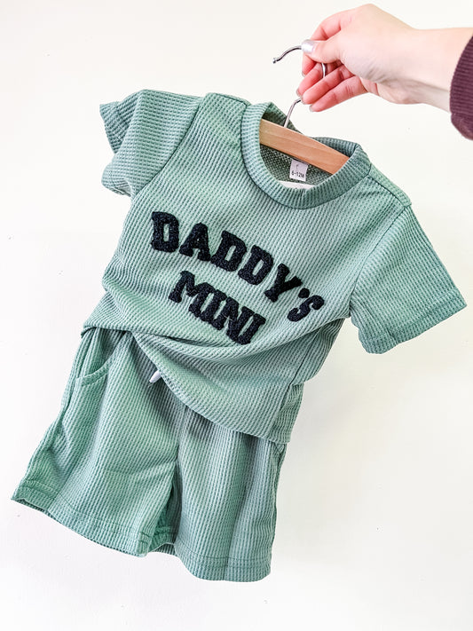 A hand displays the Tiny Tot Bundles Daddy's Mini Waffle Set—a green ribbed toddler outfit featuring a short-sleeved top with bold black "DADDYS MINI" text and matching shorts—against a plain white background.