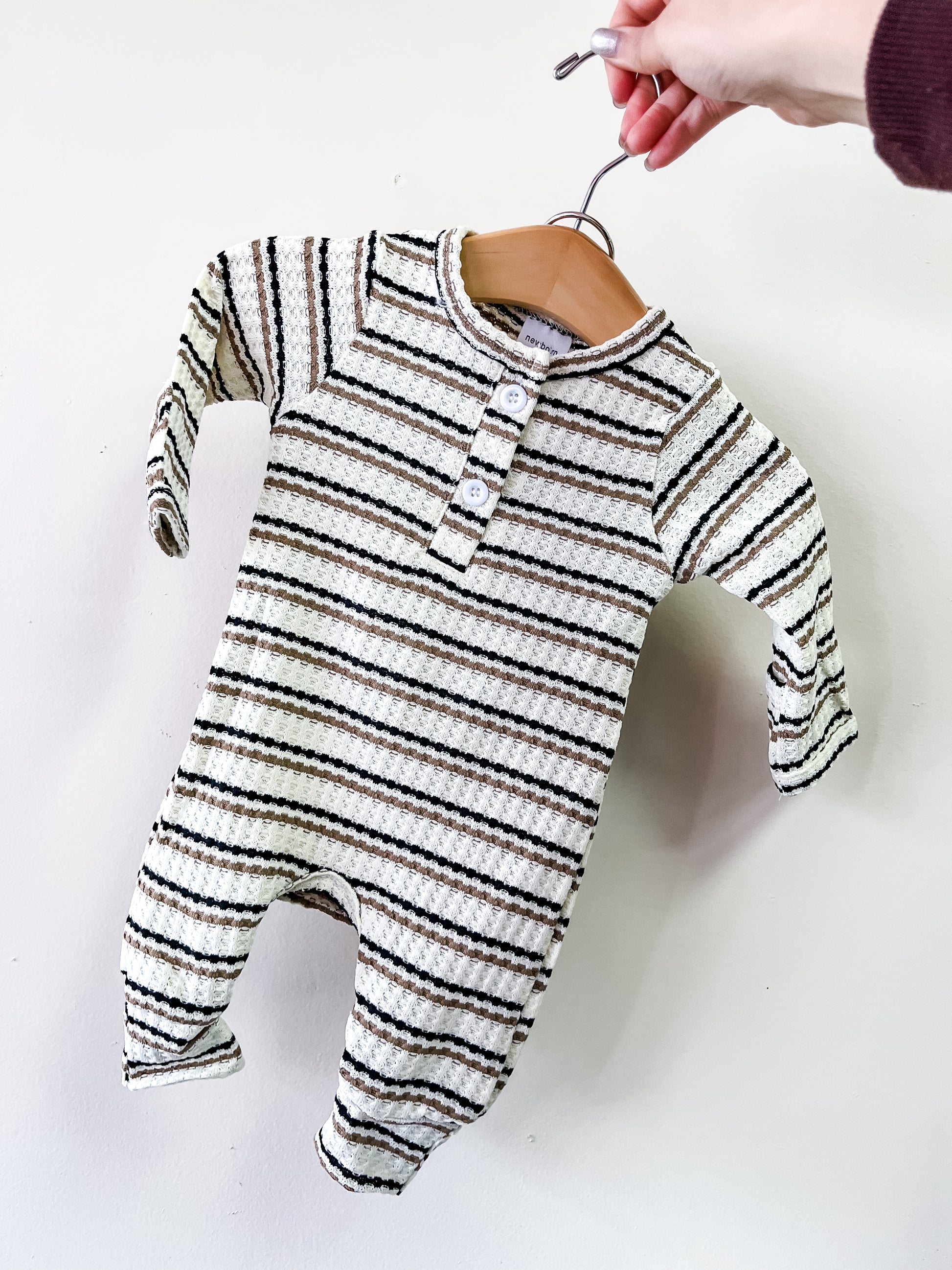 A hand holds Tiny Tot Bundles' Neutral Stripe Waffle Romper—a long-sleeved, cream baby romper with black and brown stripes and three chest buttons—against a plain white background.