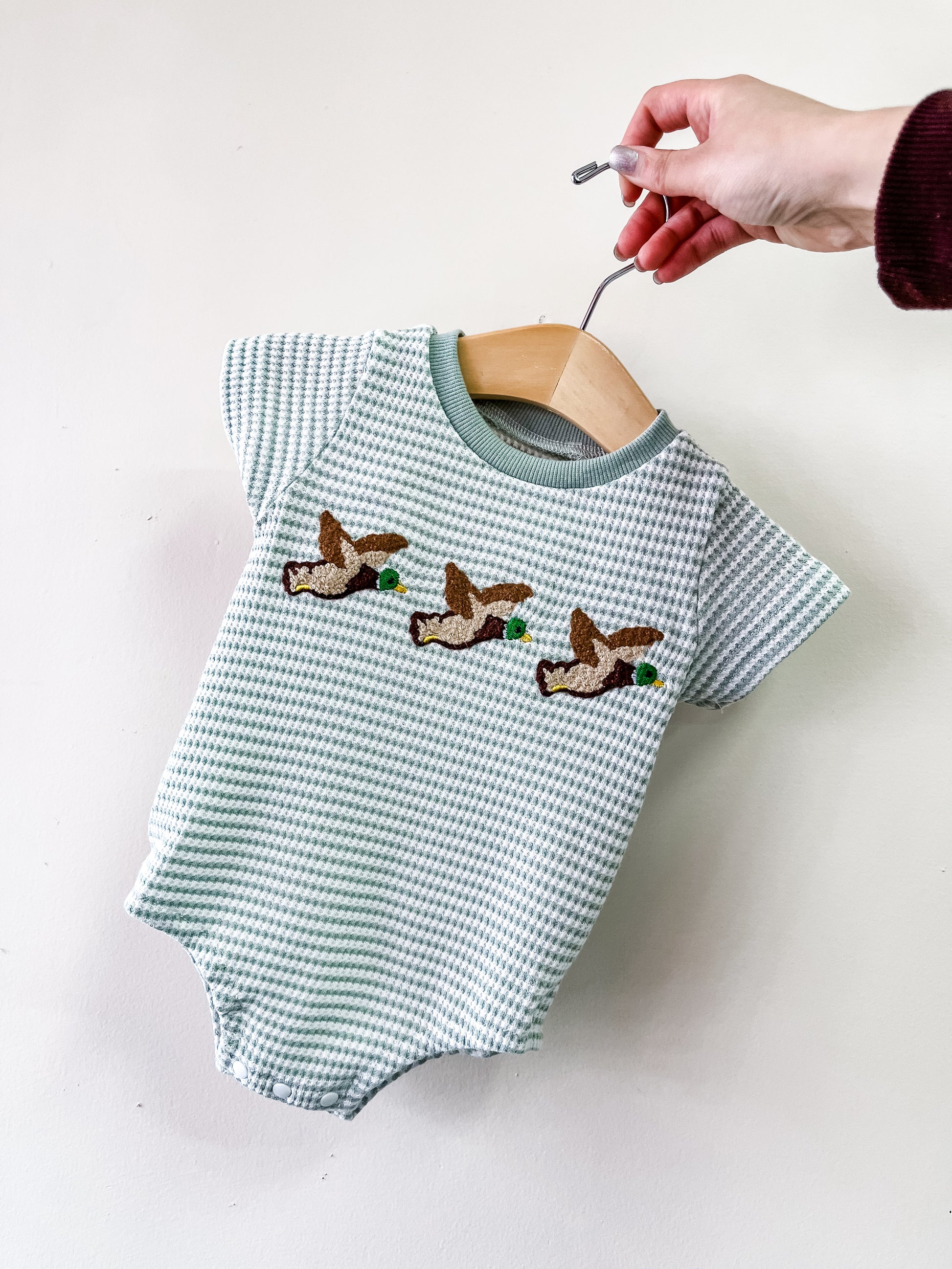 A hand holds the Tiny Tot Bundles Flying Ducks Waffle Bubble, a green and white checkered baby onesie with three embroidered flying ducks on the chest, displayed on a wooden hanger.