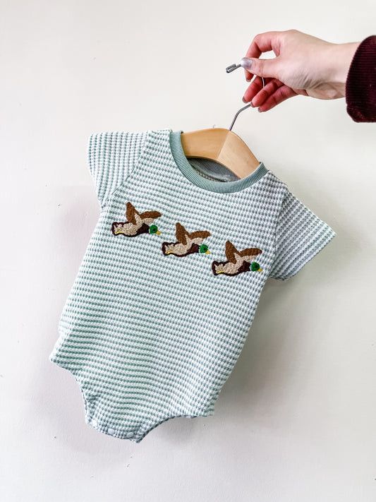 A hand holds the Tiny Tot Bundles Flying Ducks Waffle Bubble, a green and white checkered baby onesie with three embroidered flying ducks on the chest, displayed on a wooden hanger.