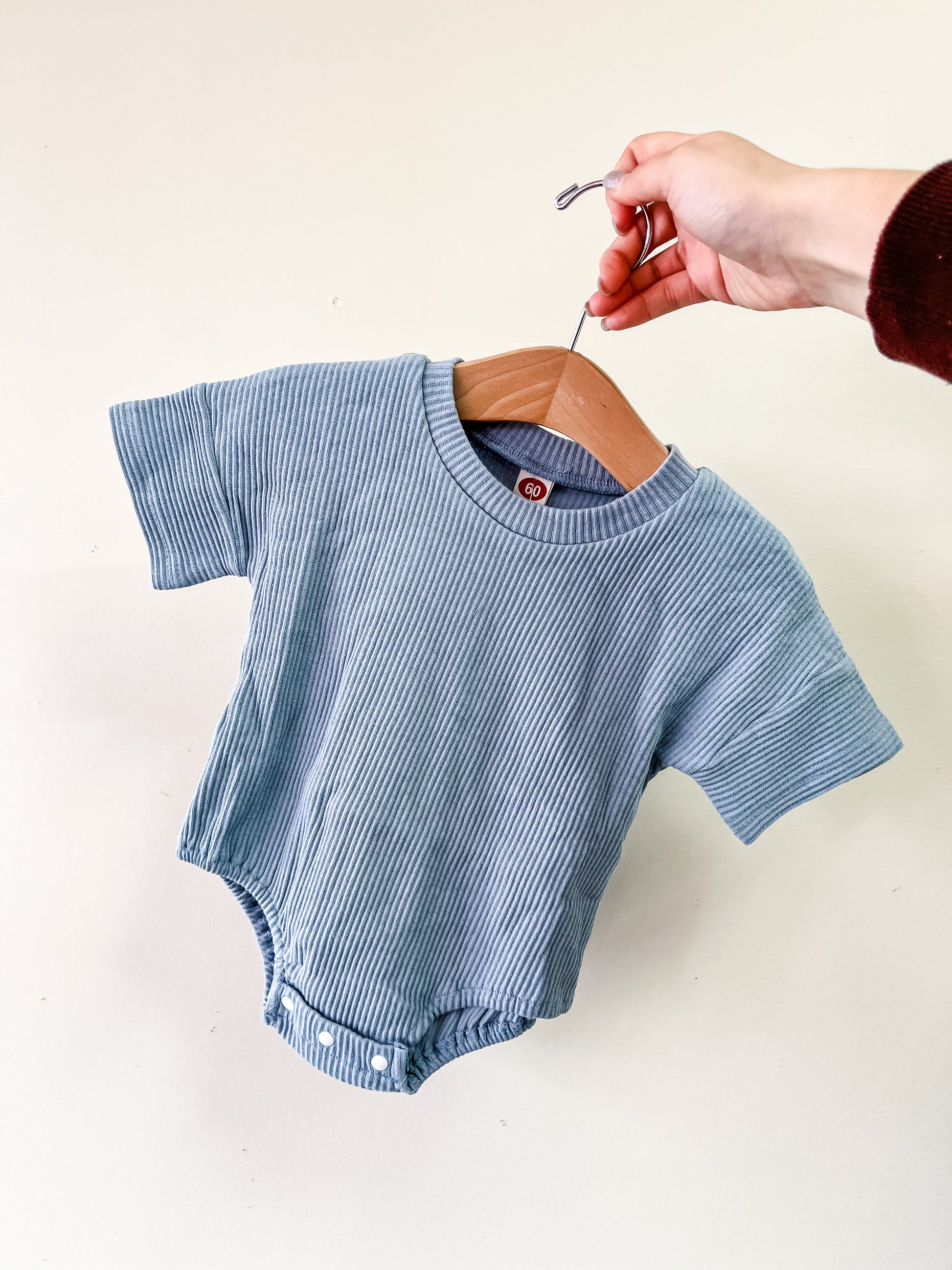 A hand holds a wooden hanger with the Tiny Tot Bundles Blue Cozy Ribbed Bubble, a short-sleeve, light blue baby onesie, against a plain light background.
