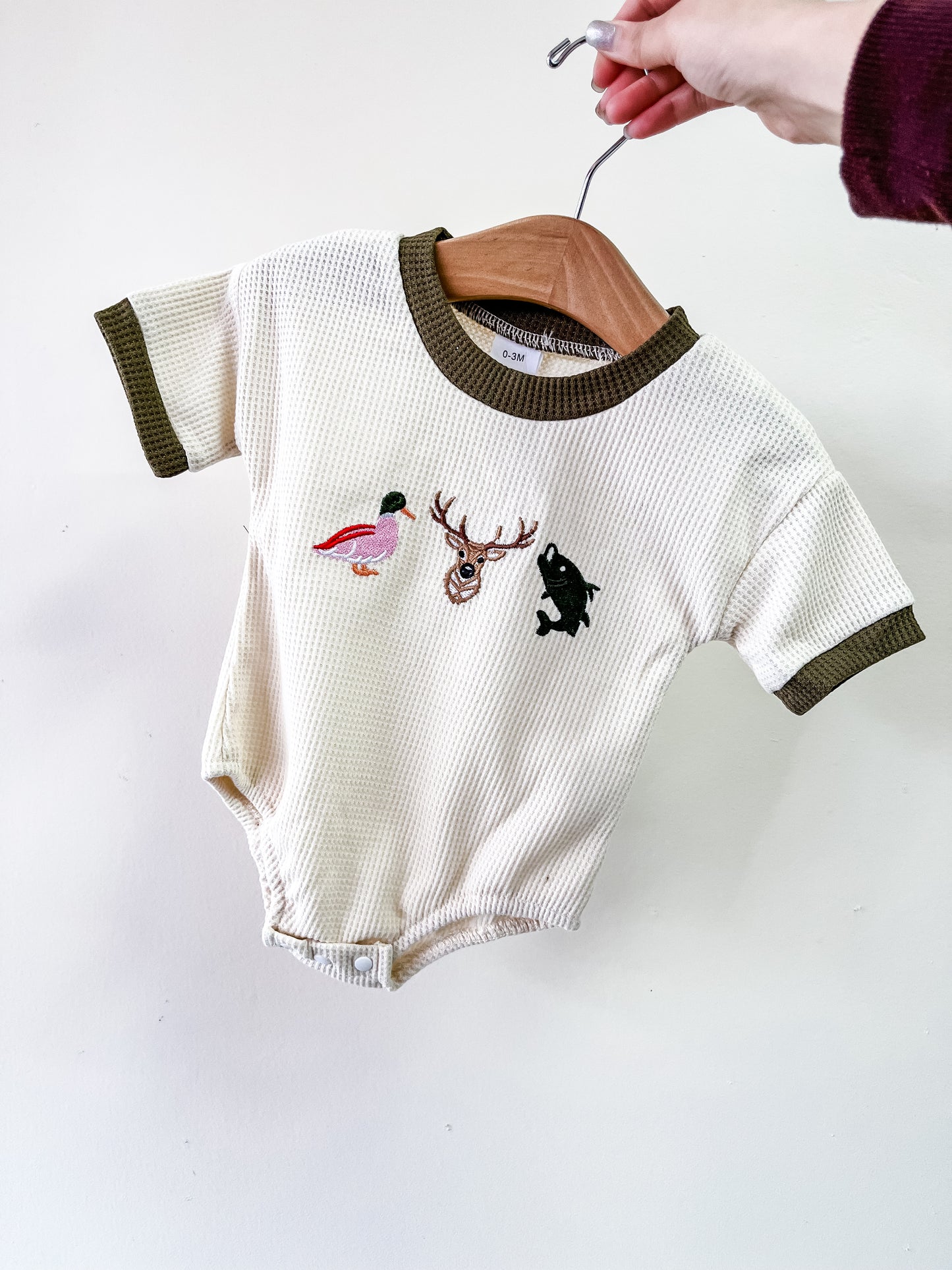 A hand holds the Tiny Tot Bundles Nature Pals Waffle Bubble, a cream baby onesie with dark green trim and small embroidered duck, deer head, and fish on the chest.