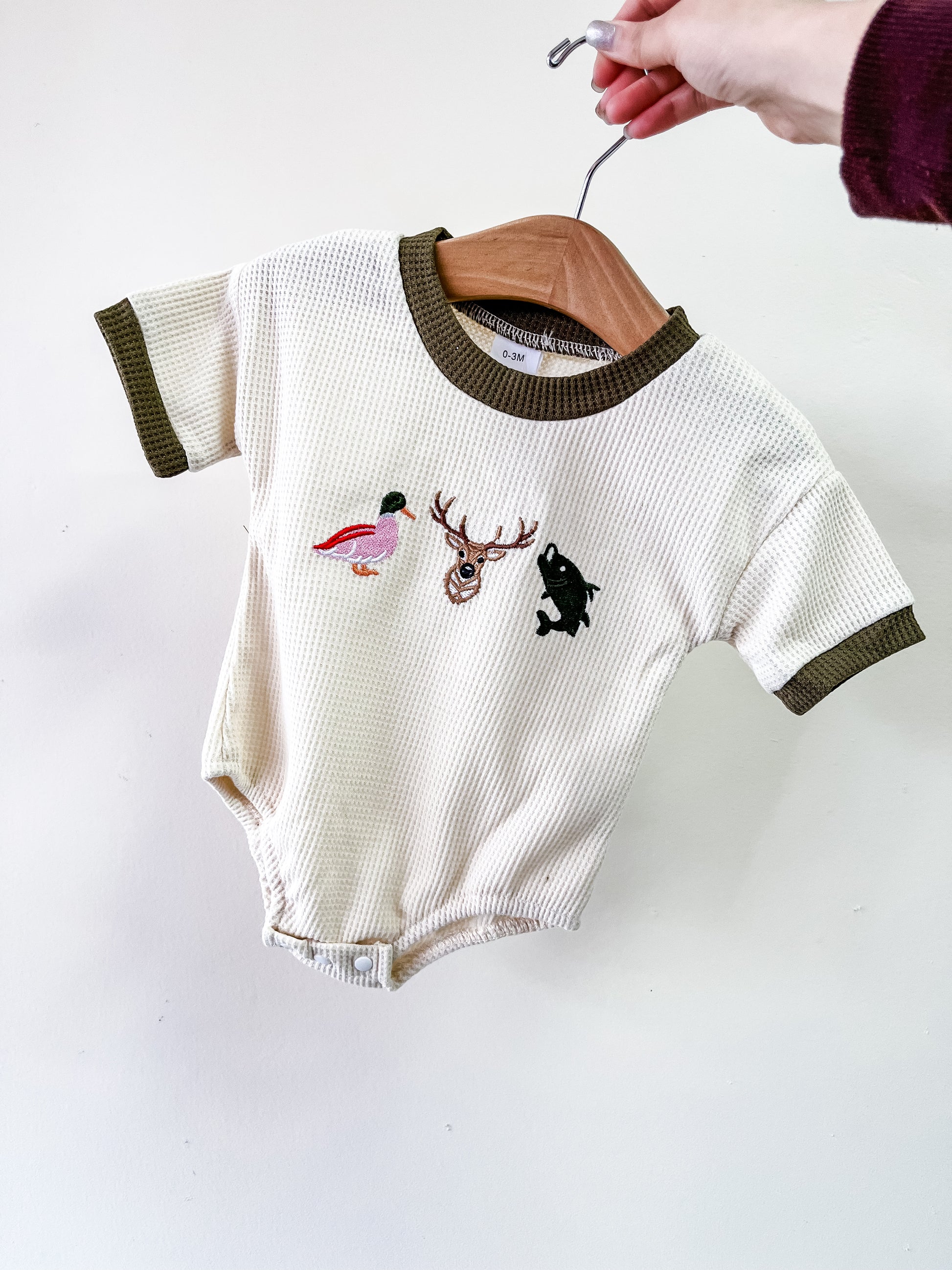 A hand holds the Tiny Tot Bundles Nature Pals Waffle Bubble, a cream baby onesie with dark green trim and small embroidered duck, deer head, and fish on the chest.