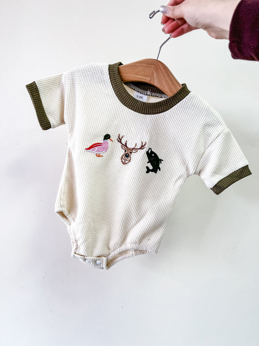 A hand holds the Tiny Tot Bundles Nature Pals Waffle Bubble, a cream baby onesie with dark green trim and small embroidered duck, deer head, and fish on the chest.