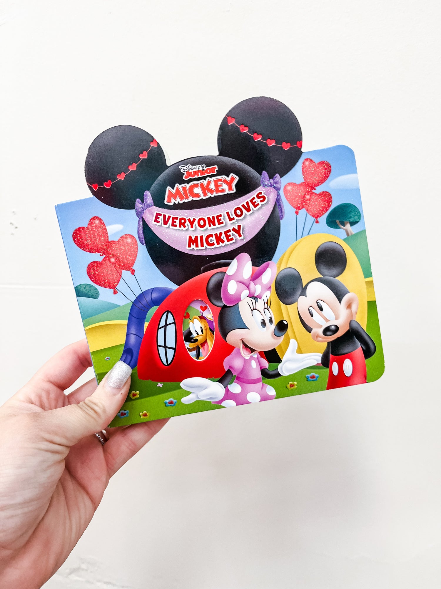 A hand holds the Tiny Tot Bundles "Everyone Loves Mickey Book," a colorful board book featuring Minnie and Mickey Mouse with heart-shaped balloons on the cover, set against cartoon scenery and a house.