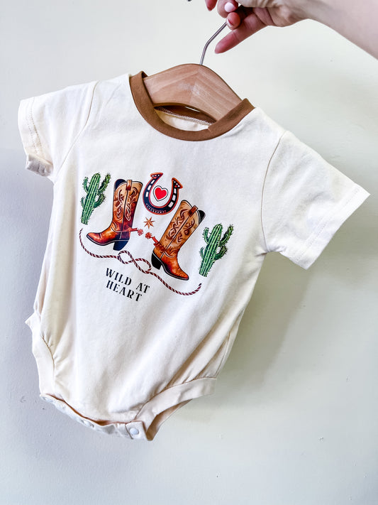 A hand holds the Tiny Tot Bundles Wild At Heart Bubble on a wooden hanger, featuring cowboy boots, cacti, a horseshoe, and “Wild at Heart” on the front.