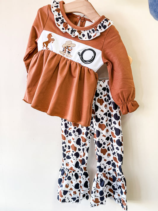 The Howdy Rodeo Babe Set by Tiny Tot Bundles features a rust long-sleeve top with a ruffled collar and Western prints, paired with white ruffled pants in a brown and black cow print. Outfit is displayed on a hanger.