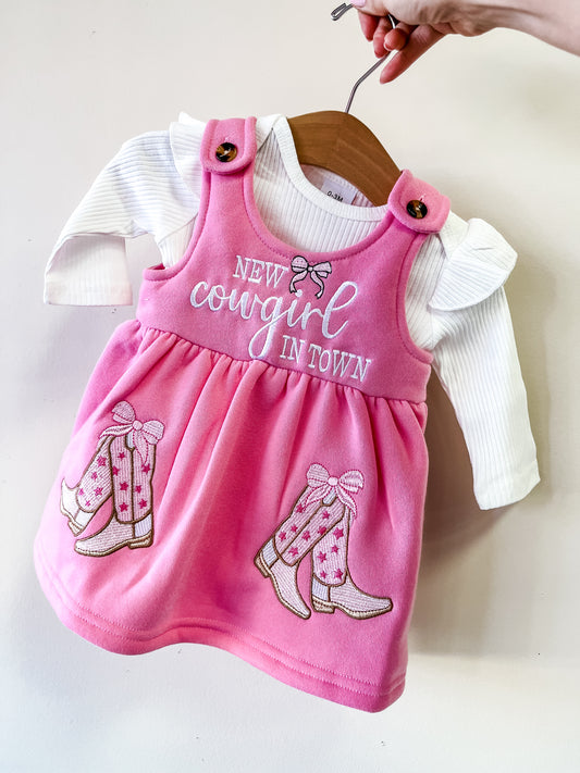 A hand holds up the Tiny Tot Bundles New Cowgirl in Town Set, featuring a pink baby dress with "New cowgirl in town" and embroidered cowboy boots, layered over a white long-sleeve shirt with ruffled shoulders.