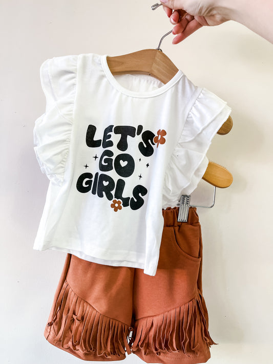 A hand holds the Let's Go Girls Ruffle Set by Tiny Tot Bundles: a kids outfit with a white ruffled-sleeve top featuring "LETS GO GIRLS" and orange flowers, paired with rust-colored fringe shorts.
