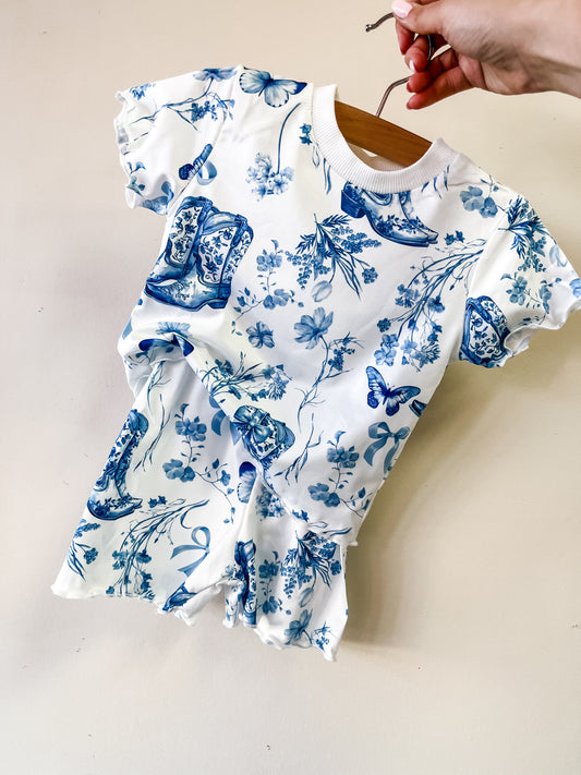 A hand holds up the Tiny Tot Bundles Bluebell Toile Set, a white baby romper with blue floral, butterfly, and shoe patterns, short sleeves, and ruffled edges. The romper is displayed on a wooden hanger against a plain light wall.