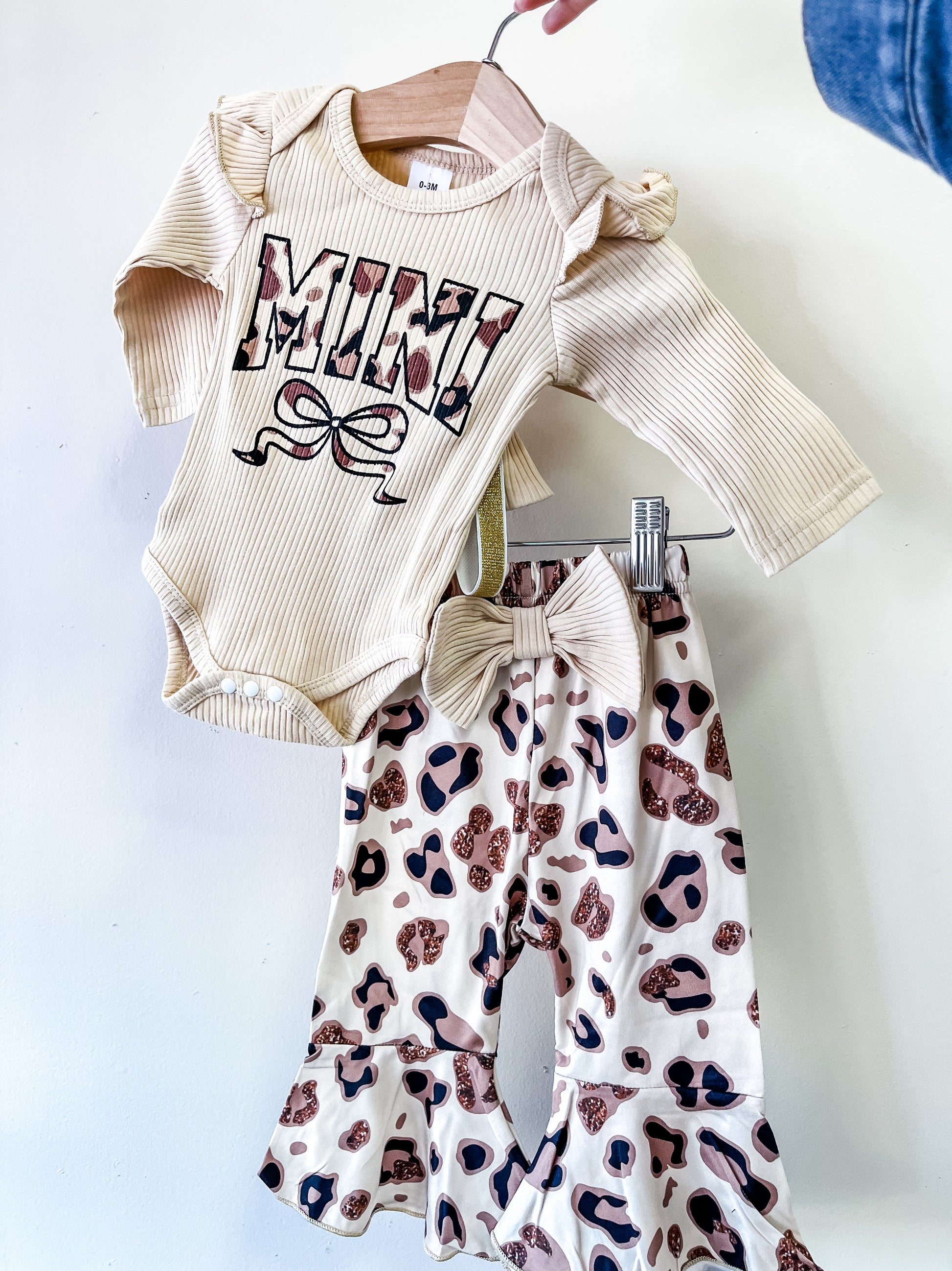 The Leopard Bow Mini Set by Tiny Tot Bundles features a cream long-sleeve onesie with "MINI" in bold, patterned letters, paired with ruffle-bottom pants in a brown and pink leopard print.