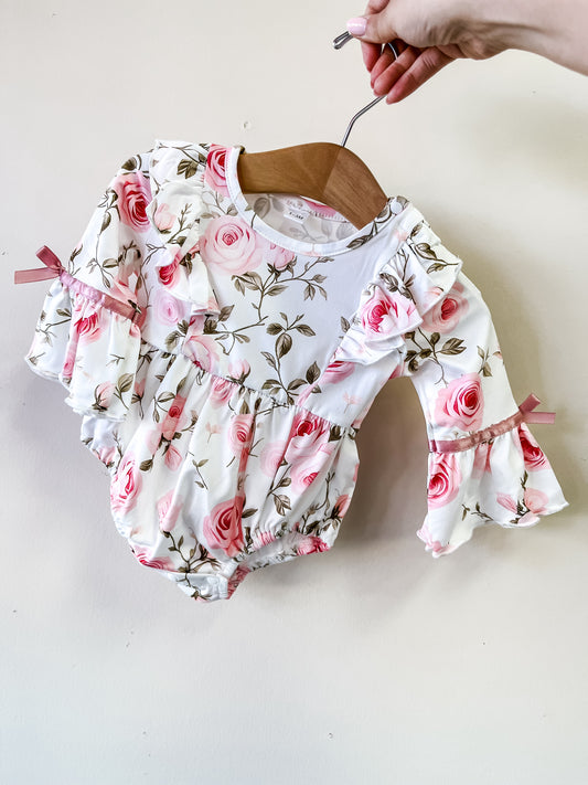 A hand holds Tiny Tot Bundles' Blushing Rose Romper, featuring a white base with pink rose florals, long ruffled sleeves, and pink ribbon bows.
