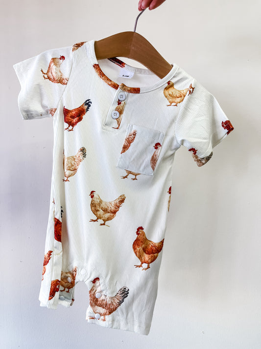 The Tiny Tot Bundles Farmyard Hen Romper is a white, short-sleeved baby romper with brown and tan chicken prints, a chest pocket, and three buttons, displayed on a wooden hanger against a plain background.