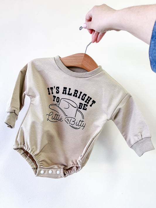 A hand holds the Little Bitty Romper by Tiny Tot Bundles, a beige long-sleeve baby onesie. The front features black text reading "ITS ALRIGHT TO BE Little Bitty" with a small cowboy hat illustration.