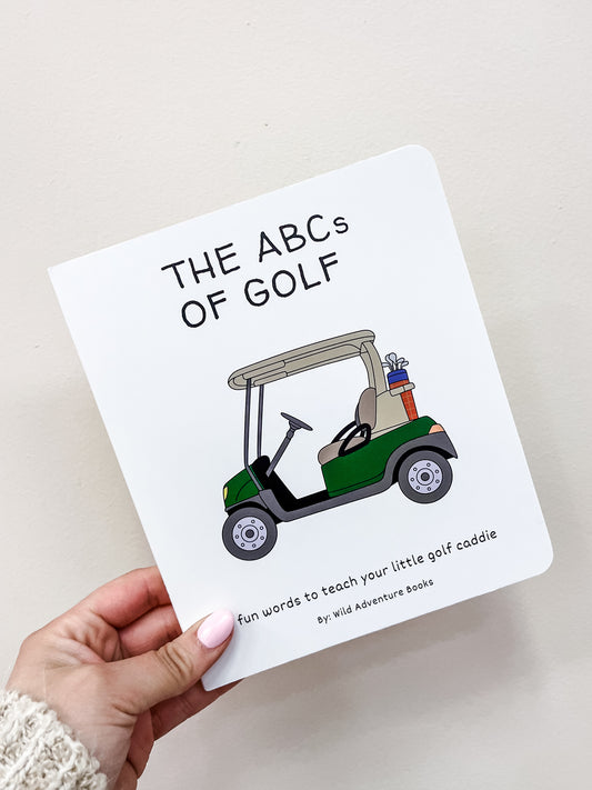 A hand holds Wild Adventure Books' "Abc's of Golf Book," showing a green golf cart with clubs on the cover and the subtitle, "Fun words to teach your little golf caddie.