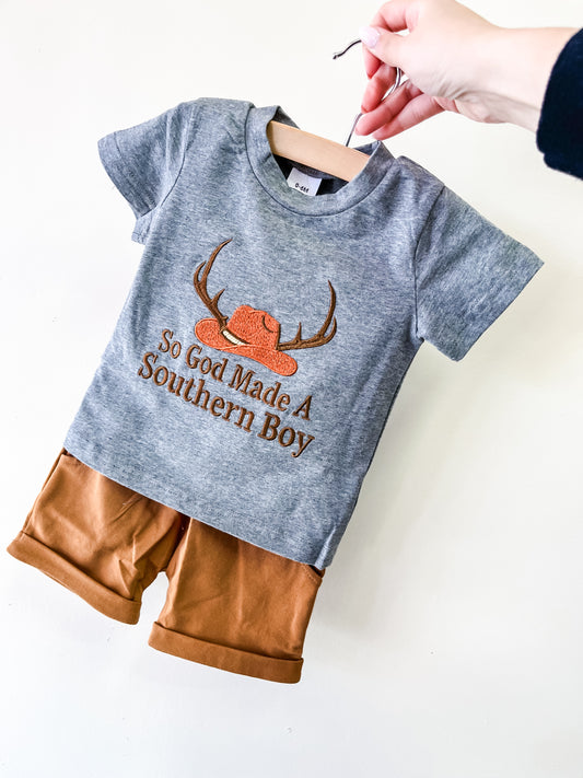 A hand holds Tiny Tot Bundles' Southern Blessings Short Set, featuring a gray “So God Made A Southern Boy” t-shirt with a hat and antlers graphic, and matching brown shorts, displayed against a plain white background.