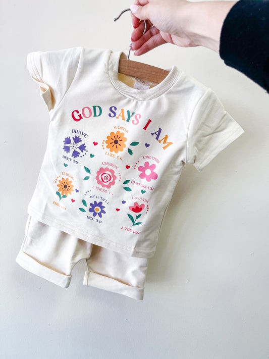 A hand holds a hanger with the Sunday Blessing Blue Set by Tiny Tot Bundles, a toddler outfit. The shirt displays "God Says I Am" with colorful flowers and uplifting words like Brave, Chosen, and Cherished.