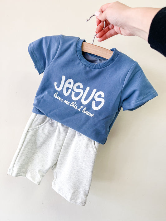 A hand displays the Jesus Loves Me Blue Set by Tiny Tot Bundles: a blue baby t-shirt with "JESUS loves me this I know" in white letters, paired with light gray shorts, against a plain background.