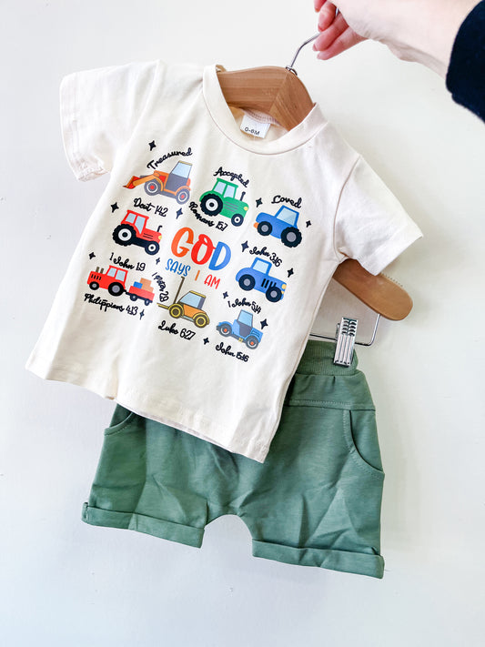 A hand holds the Tiny Tot Bundles "God Says I Am Set," showing a toddler outfit with a white T-shirt featuring colorful cars and Bible verses, plus sage green shorts, against a plain white background.