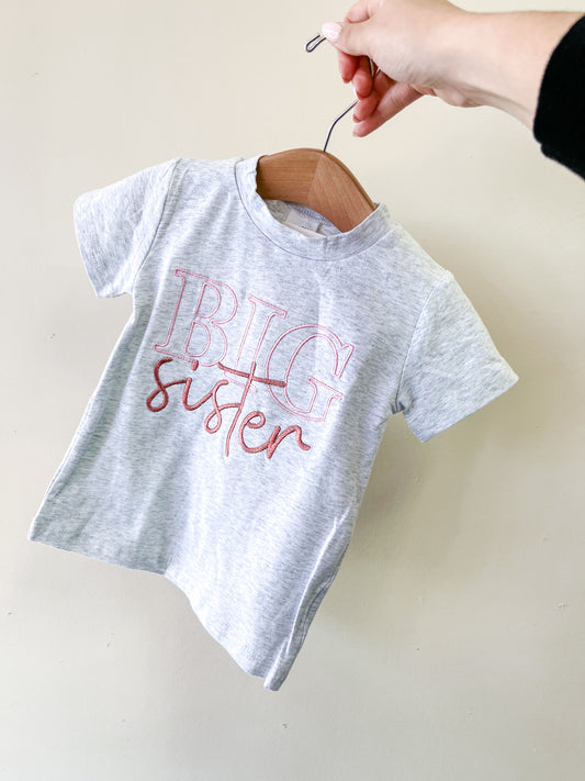 A hand holds a wooden hanger with a light gray Tiny Tot Bundles Big Sister Scripted Tee featuring "BIG sister" in bold pink and red letters, shown against a plain light background.