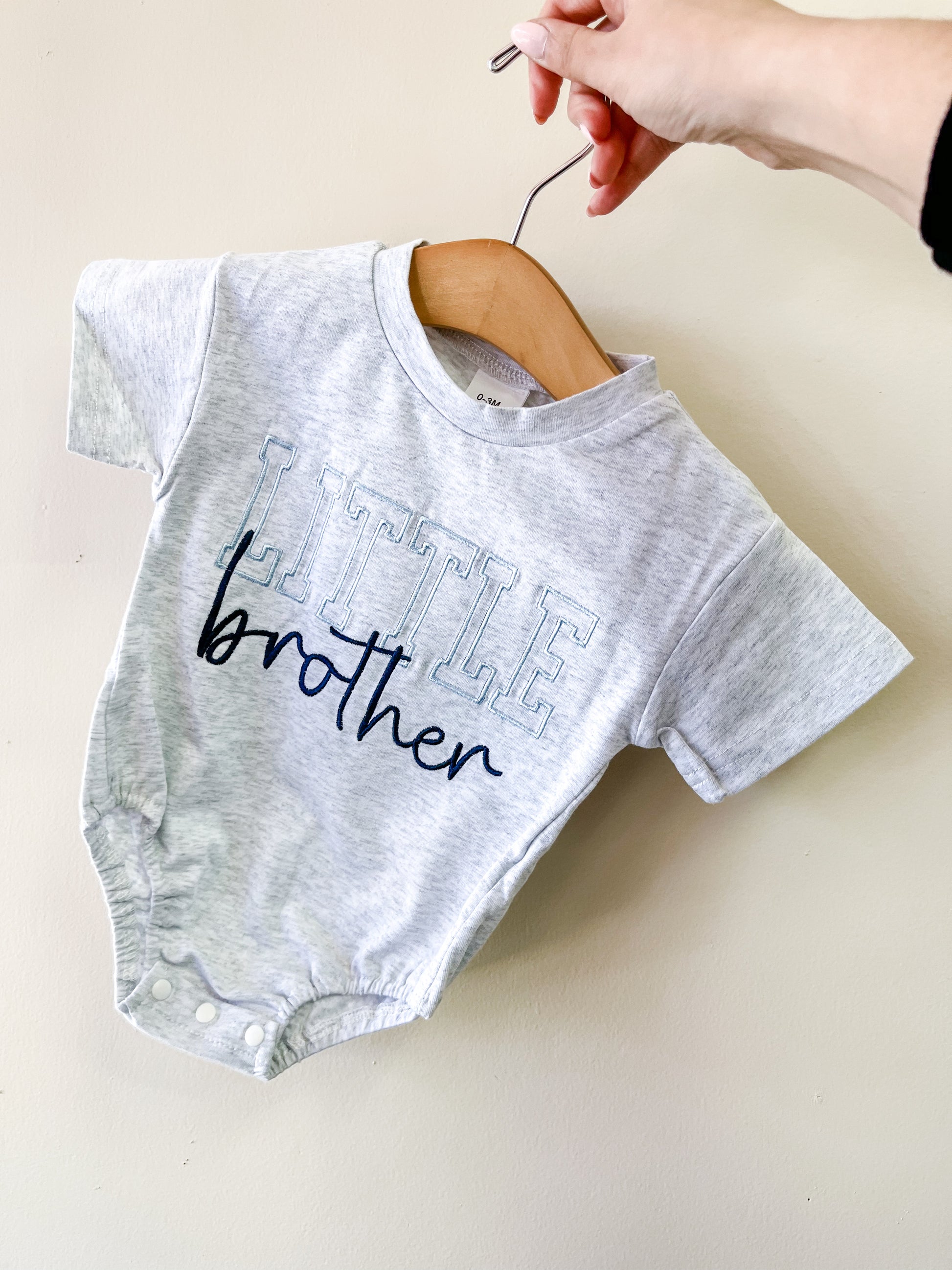 A hand holds a wooden hanger with the Tiny Tot Bundles Little Brother Scripted Bubble, a light gray baby onesie featuring "LITTLE brother" in large outlined and cursive letters on the front.