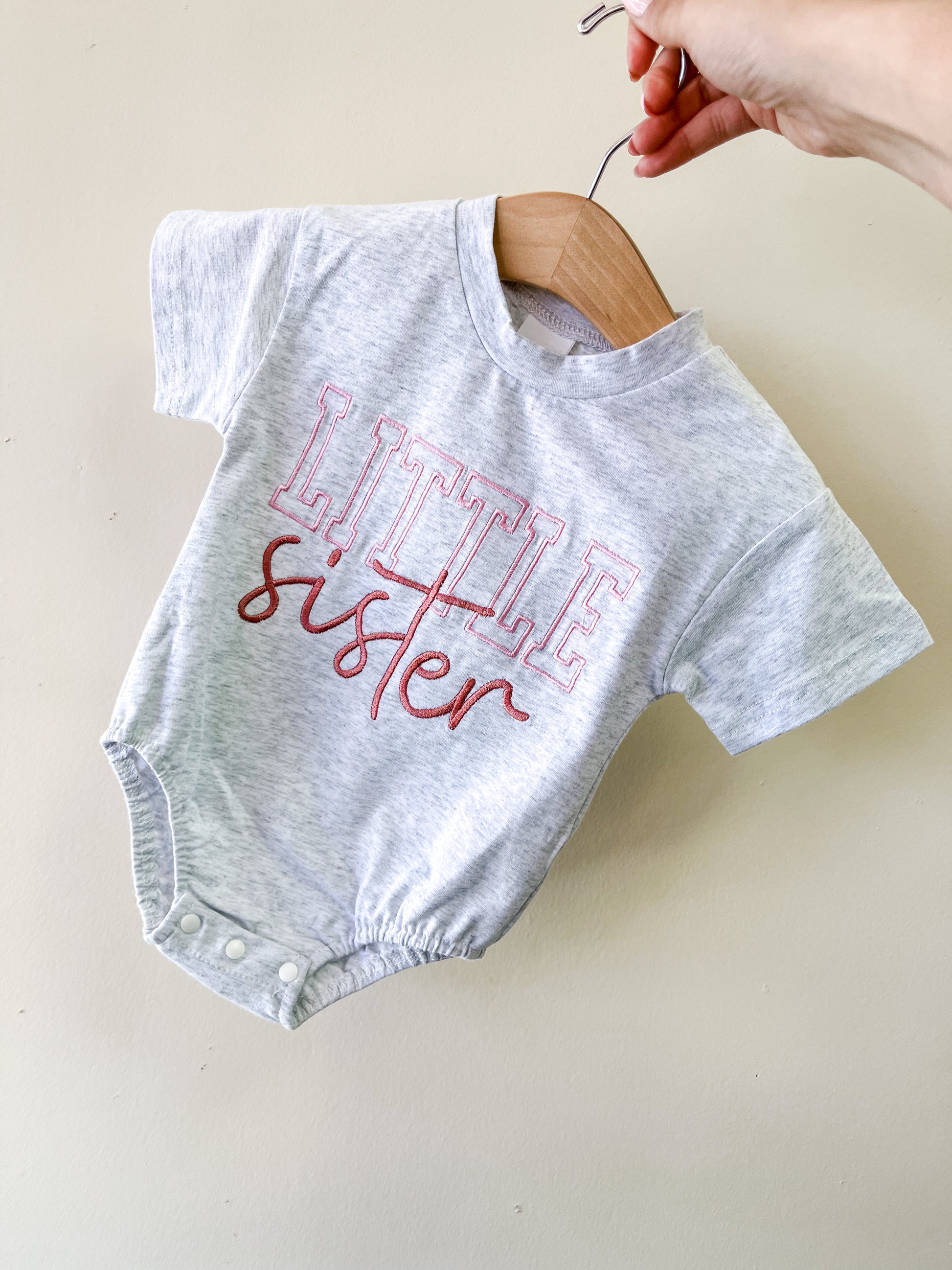 A hand holds a hanger with Tiny Tot Bundles' Little Sister Scripted Bubble, a light gray baby onesie featuring "LITTLE sister" in pink and red letters, displayed against a plain light-colored wall.