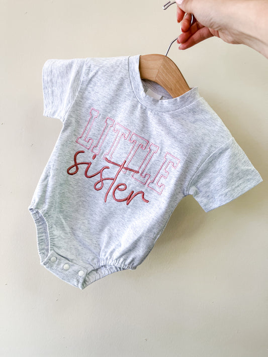 A hand holds a hanger with Tiny Tot Bundles' Little Sister Scripted Bubble, a light gray baby onesie featuring "LITTLE sister" in pink and red letters, displayed against a plain light-colored wall.