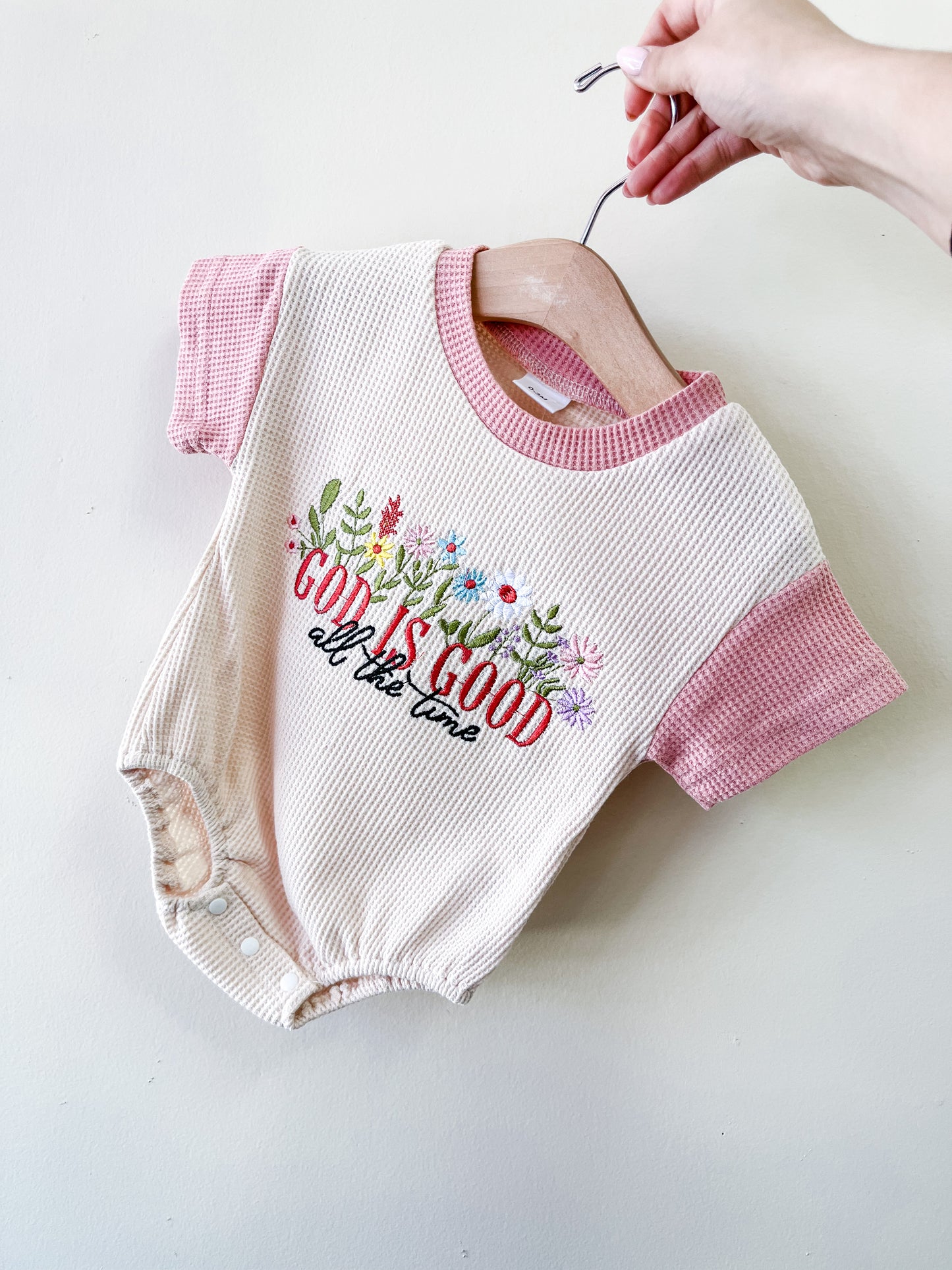 A hand holds the Blessed Blooms Bubble by Tiny Tot Bundles—a cream onesie with pink sleeves, colorful embroidered flowers, and "God is good all the time" on the front.